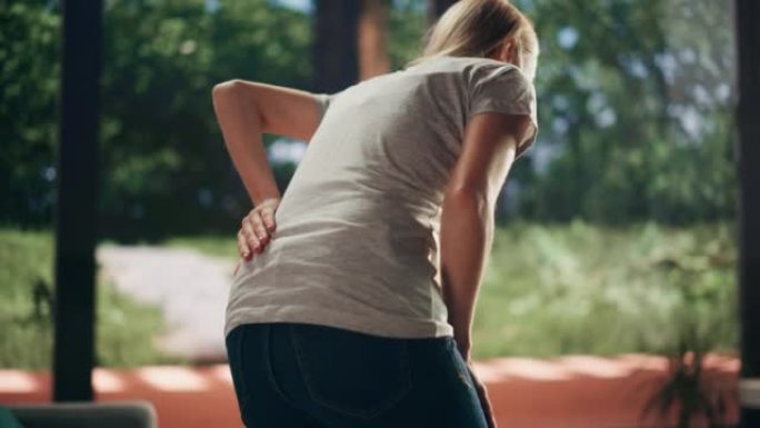 一名年轻的成年女性在家中从沙发上站起来，并因脊柱创伤而感到背部疼痛。按摩和伸展背部以减轻不适和伤害。