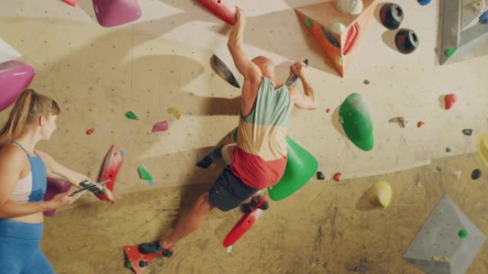 攀岩教练在健身房的抱石墙上给初学者上课。女运动员拿着平板电脑，展示正确的攀爬技术，以强健男性。