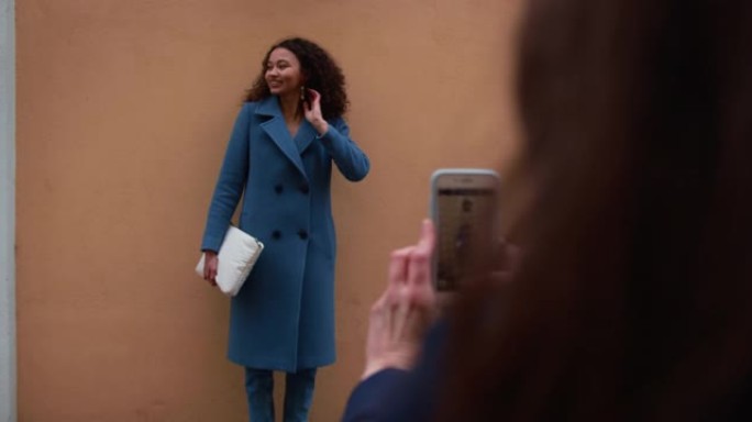 美丽的年轻快乐20-25混血女人穿着蓝色外套在墙上摆姿势，由她的朋友用智能手机拍照