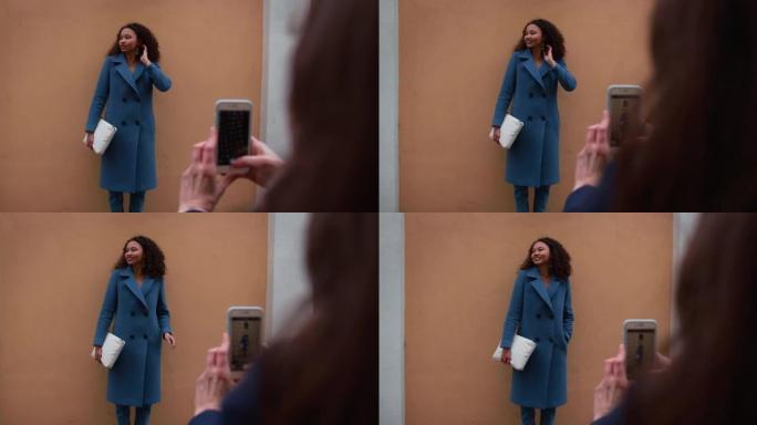 美丽的年轻快乐20-25混血女人穿着蓝色外套在墙上摆姿势，由她的朋友用智能手机拍照