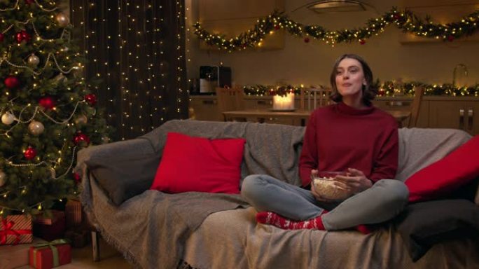 一位穿着红色毛衣的迷人棕色头发的女人正在看电视，坐在沙发上，拿着一碗爆米花，在装饰有圣诞树的节日房间