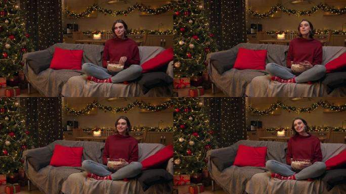 一位穿着红色毛衣的迷人棕色头发的女人正在看电视，坐在沙发上，拿着一碗爆米花，在装饰有圣诞树的节日房间