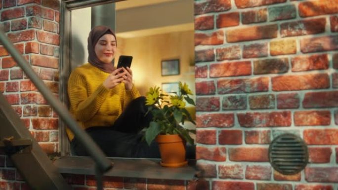 年轻的穆斯林女性坐在多层砖房的窗台上时使用智能手机。富有创造力的女性在社交媒体上与朋友联系时微笑并感