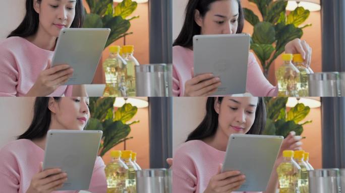 美丽的亚洲年轻女子年龄30岁，穿着粉红色衬衫坐在沙发上，在家中通过平板电脑检查消费品。在线购物概念。
