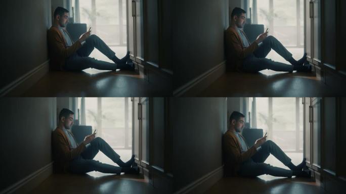 年轻成年男子在家中使用智能手机的肖像。他坐在走廊地板上，背靠墙。使用互联网检查网络和聊天，在社交媒体