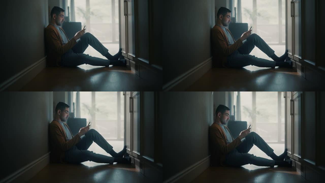 年轻成年男子在家中使用智能手机的肖像。他坐在走廊地板上，背靠墙。使用互联网检查网络和聊天，在社交媒体