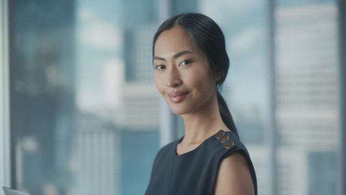 美丽的亚洲女商人肖像，穿着时髦的黑色连衣裙，在摩天大楼的大城市办公室的窗户旁边摆姿势。自信的女CEO