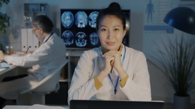 迷人的亚洲女医生微笑的慢动作肖像坐在办公桌前，笔记本电脑在黑暗的办公室里