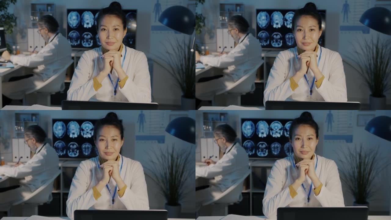 迷人的亚洲女医生微笑的慢动作肖像坐在办公桌前，笔记本电脑在黑暗的办公室里