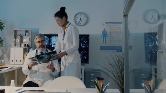 亚洲女医生走在黑暗的医院办公室，与男性同事白人男子交谈，看着核磁共振扫描
