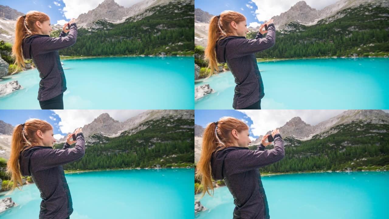 绿松石山湖的女游客欣赏美景，用智能手机拍照，背景中的山脉