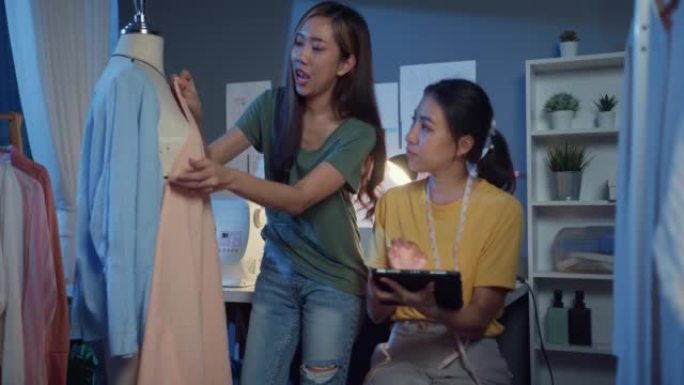 两位专业的亚洲开朗女士时装设计师团队合作，休闲加班，举办平板电脑分享想法，晚上在商店假人身上创造漂亮