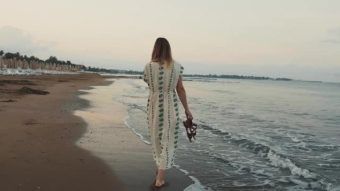 镜头跟随年轻快乐的女人穿着漂亮的衣服，赤脚沿着温暖的黄昏热带沙滩在海浪边缘行走。