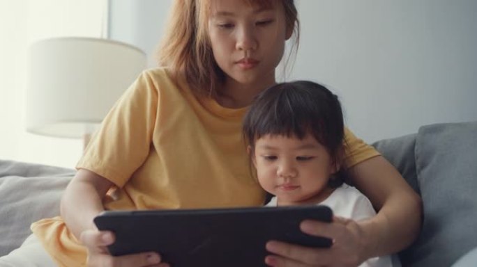 快乐快乐的亚洲家庭妈妈和可爱的孩子使用数字平板电脑兴趣卡通，看电影玩得开心在家里客厅的沙发上放松。
