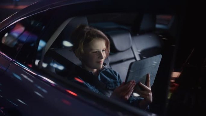 小男孩坐在汽车后座上，晚上通勤回家。乘客在平板电脑上观看有趣的娱乐活动，同时在城市街道上乘坐出租车，