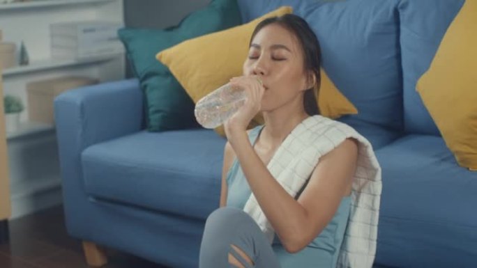 微笑的年轻迷人的运动亚洲强壮的女人，用毛巾喝水在家里的客厅锻炼后休息一下。家庭检疫锻炼和健身健康的生