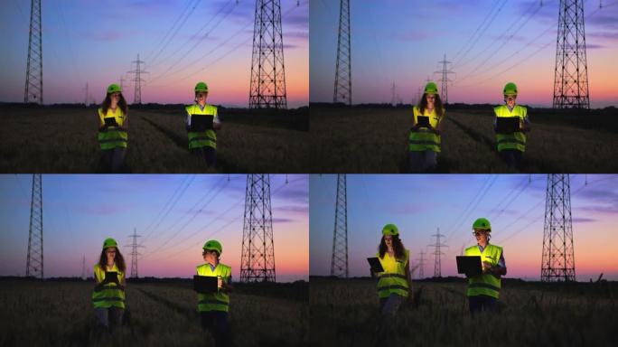 慢动作，两名电气工程师看着平板电脑和笔记本电脑的跟踪镜头，高架电力线和背景中的日落