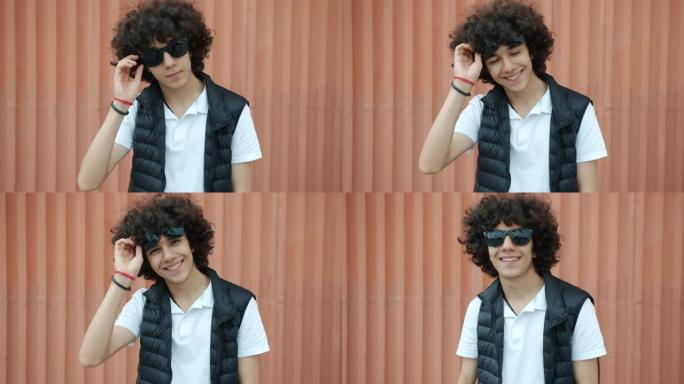 时尚的中东少年少年举着太阳镜在户外微笑的慢动作肖像