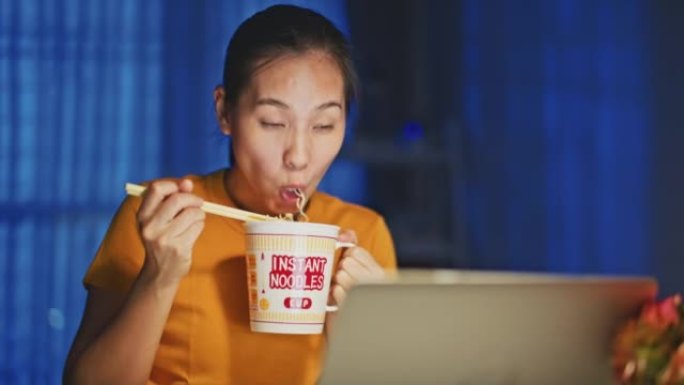 亚洲女子晚上在家用笔记本电脑工作时吃方便面