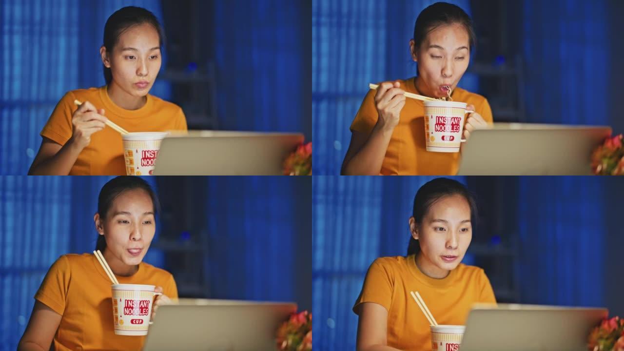 亚洲女子晚上在家用笔记本电脑工作时吃方便面