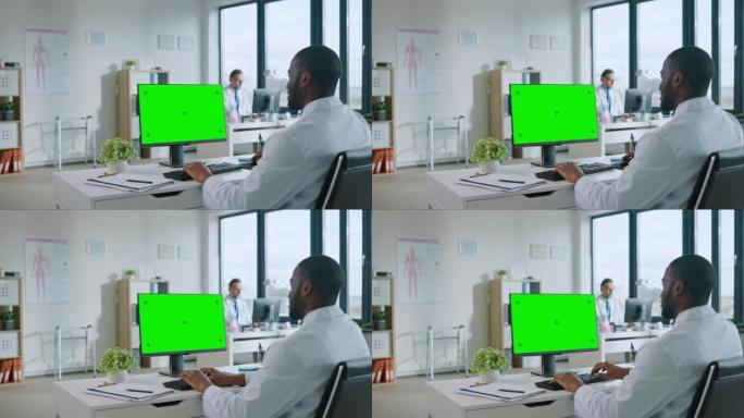黑人医生正在健康诊所的绿屏模拟显示计算机上工作。穿着白大褂的助理正在医院办公室的桌子后面阅读病史。
