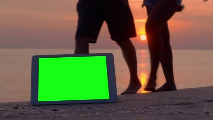 绿屏平板电脑和海滩上的跟踪点，而游客在度假背景下在海上的红色日落中行走。新技术概念的绿屏。