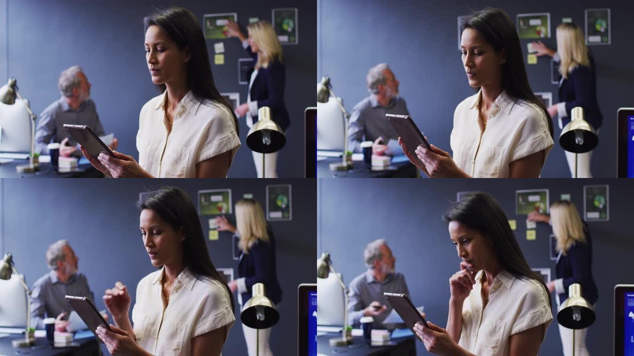 混血女商人在办公室使用数字平板电脑，同事在背后讨论