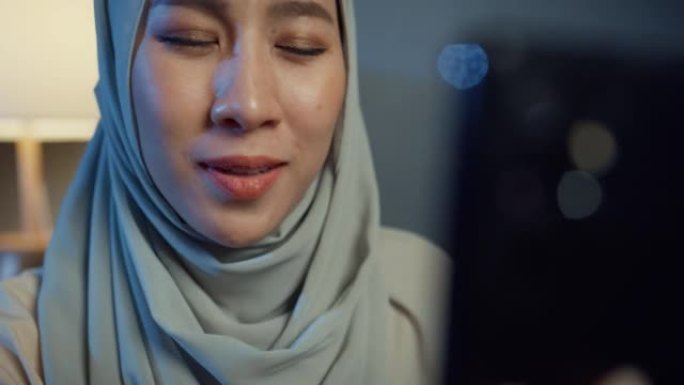特写迷人的快乐亚洲穆斯林妇女戴着头巾，随意坐在沙发沙发上，晚上在家里的客厅和男朋友一起使用智能手机视