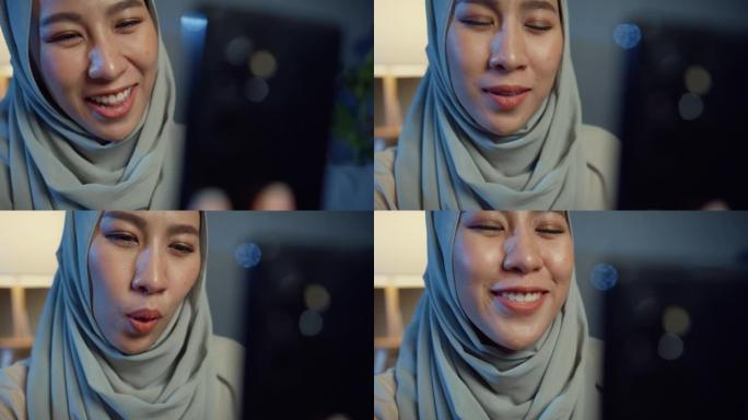 特写迷人的快乐亚洲穆斯林妇女戴着头巾，随意坐在沙发沙发上，晚上在家里的客厅和男朋友一起使用智能手机视