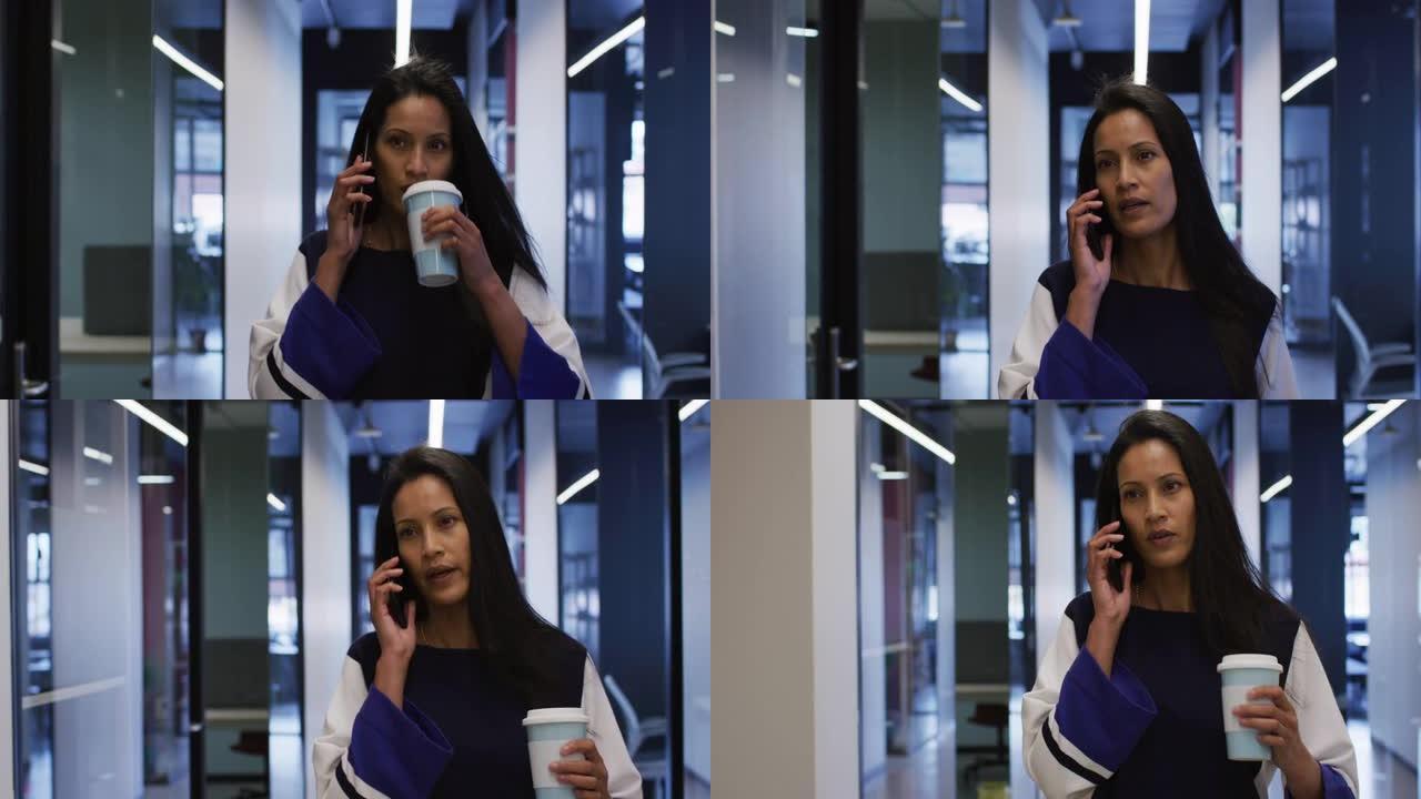 混血女商人在智能手机上聊天，拿着咖啡走在办公室走廊上
