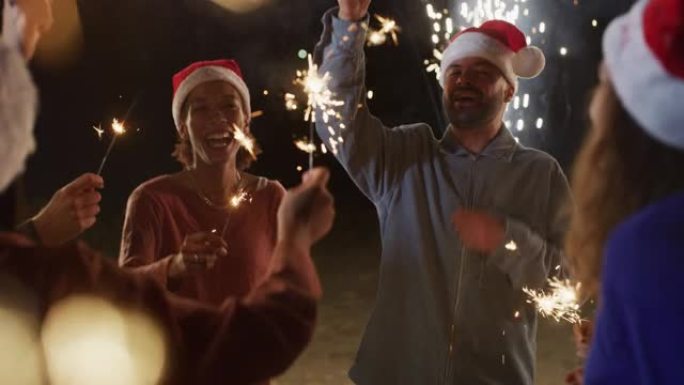 电影拍摄的是年轻的快乐微笑的多民族朋友，手里拿着圣诞老人的帽子和烟火，他们在晚上的户外庆祝晚会上唱歌