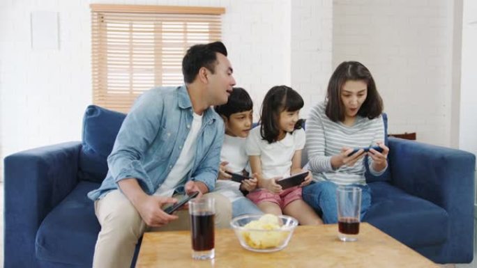 快乐的亚洲家庭爸爸，妈妈和孩子们在智能手机上在线玩有趣的游戏，坐在家里的客厅沙发上。