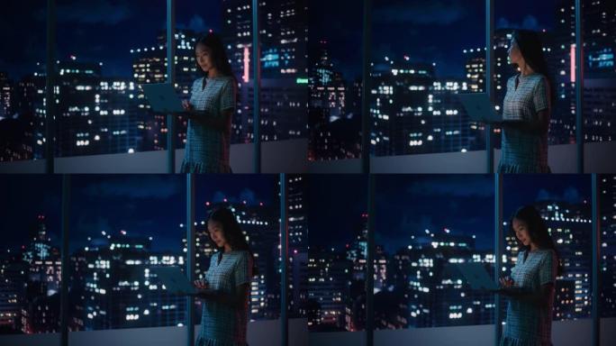 金融分析师使用笔记本电脑，站在窗户旁边，美丽的夜晚城市天际线与摩天大楼。自信的亚洲女商人在现代公司办