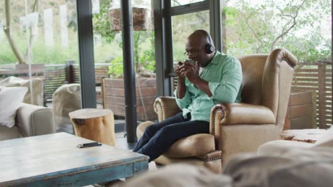 快乐的非洲裔美国老人坐在扶手椅上放松，戴着耳机喝咖啡