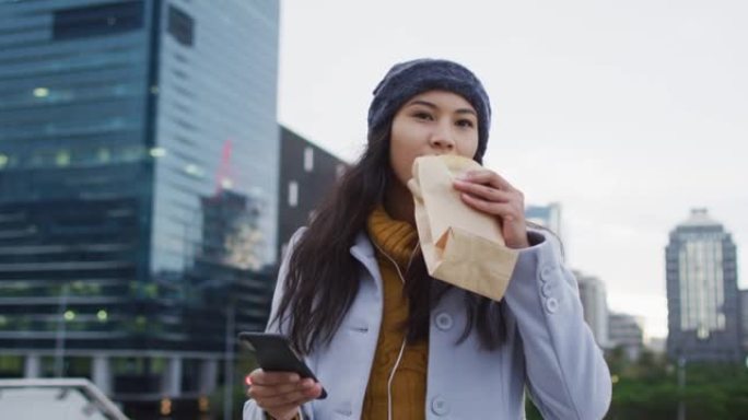 亚洲妇女使用智能手机走路并吃三明治