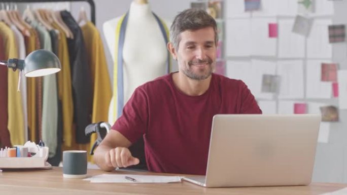 成熟的男性时装设计师坐在工作室的轮椅上，在笔记本电脑上工作，喝咖啡