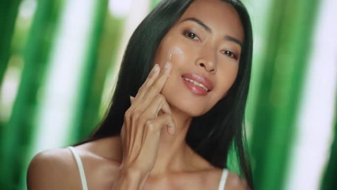 美丽的亚洲女人轻轻涂抹面霜的肖像。年轻的成年女性使用科学先进的天然化妆品护肤品使皮肤柔软光滑。绿色森