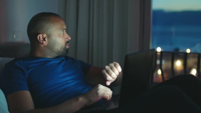 疲倦的人在晚上用笔记本电脑工作，而外面很暗，却完成了关闭计算机的工作。非裔美国人在床上工作在线，检查