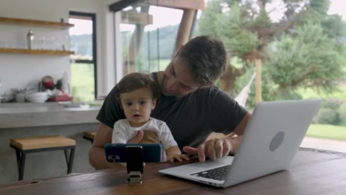 年轻英俊的父亲在笔记本电脑上工作，同时背着正在智能手机上观看视频的蹒跚学步的孩子