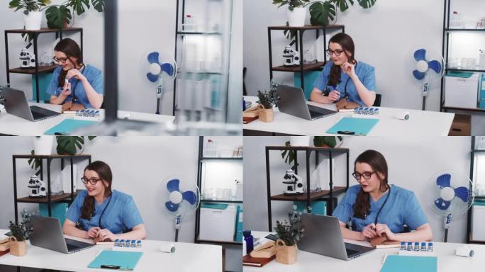 年轻的女药房医生穿着蓝色制服，通过笔记本电脑网络摄像头在线咨询远程患者，展示疫苗烧瓶。
