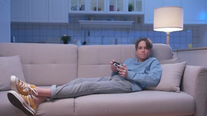 年轻英俊的游戏男孩坐在沙发上，在数字控制台上玩视频游戏的成功。兴奋的玩家控制视频游戏操纵杆。一个人专