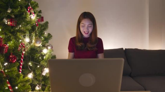 年轻的亚洲女性使用笔记本电脑视频通话与圣诞礼物盒子的情侣交谈，圣诞树装饰着家里客厅的装饰品。