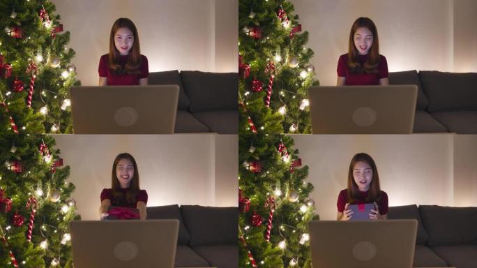 年轻的亚洲女性使用笔记本电脑视频通话与圣诞礼物盒子的情侣交谈，圣诞树装饰着家里客厅的装饰品。