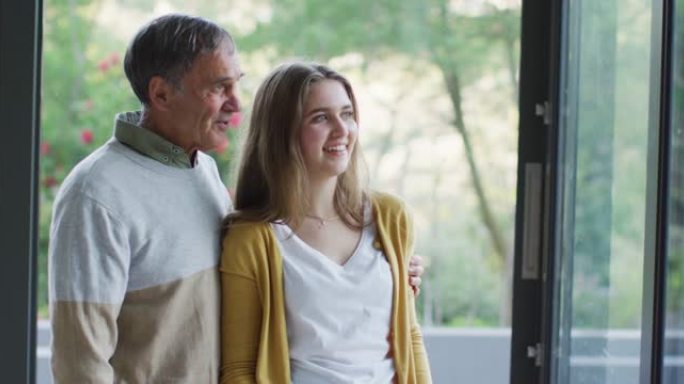 微笑的白人高级父亲和十几岁的女儿拥抱并看着窗外