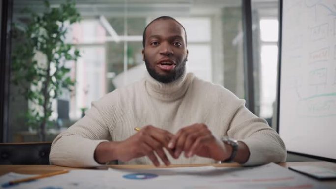 从镜头的角度来看，英俊的年轻黑人项目经理在创意办公环境中进行视频通话。快乐的非洲裔美国男性专家通过实