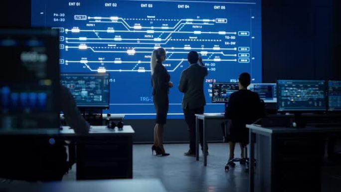 项目经理和计算机科学工程师在使用大屏幕显示基础设施信息图表和数据时交谈。电信公司系统控制和监控室