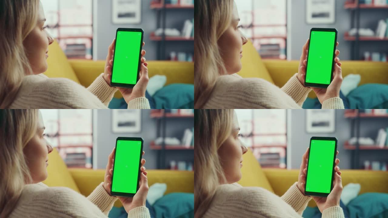 美丽的正宗女性躺在沙发上，在家里的客厅使用带有绿屏模拟显示的智能手机。她正在浏览互联网并在社交网络上