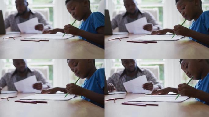 非裔美国儿子坐在厨房的桌子上做学校工作，父亲在后台工作