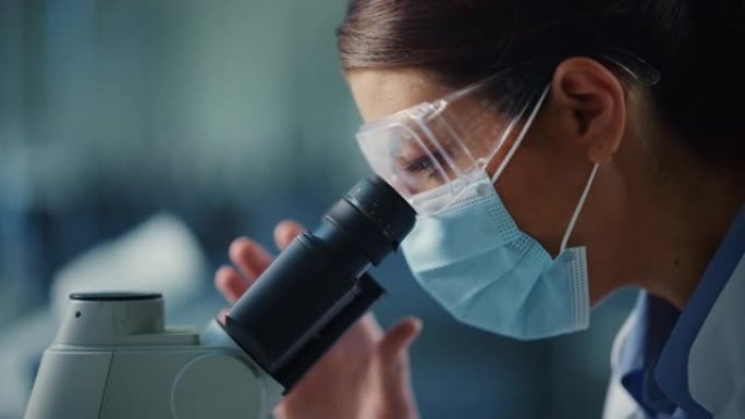 宏观特写一位戴着口罩和护目镜的女科学家在显微镜下观察的镜头。用技术设备在现代实验室中研究分子样品的女