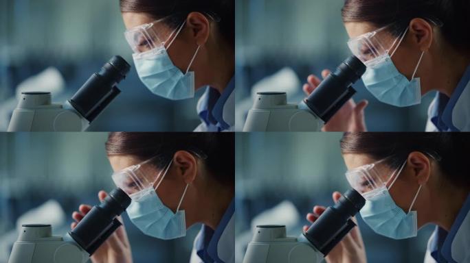 宏观特写一位戴着口罩和护目镜的女科学家在显微镜下观察的镜头。用技术设备在现代实验室中研究分子样品的女